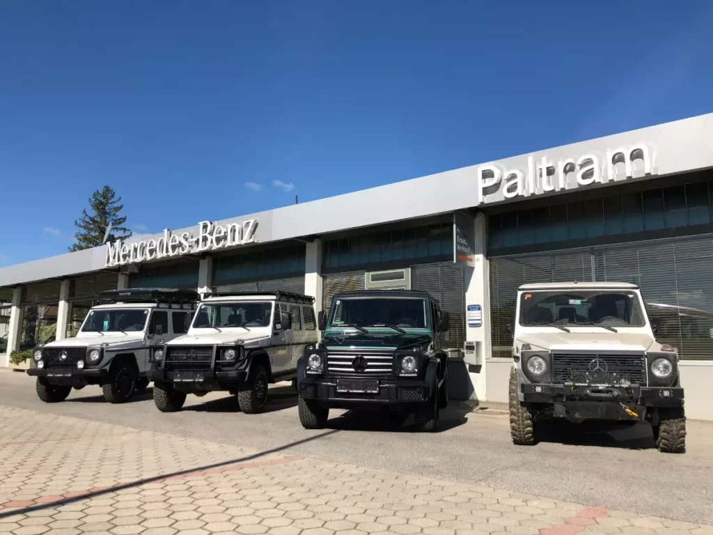 drei weiße und ein dunkelgrüner Mercedes Benz vor dem Autohaus bei Sonnenschein und blauem Himmel